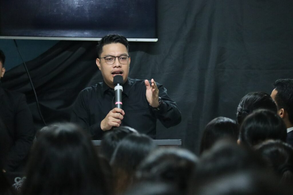 man in black long sleeve shirt holding microphone in front of and gesturing to connect with the crowd.